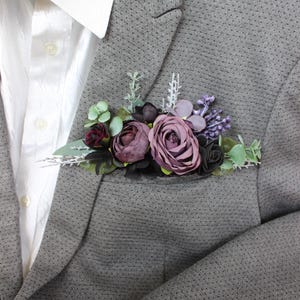 May include: A gray blazer with a floral pocket square featuring burgundy, purple, and green artificial flowers. The blazer is paired with a white dress shirt. The pocket square includes roses and other floral elements.