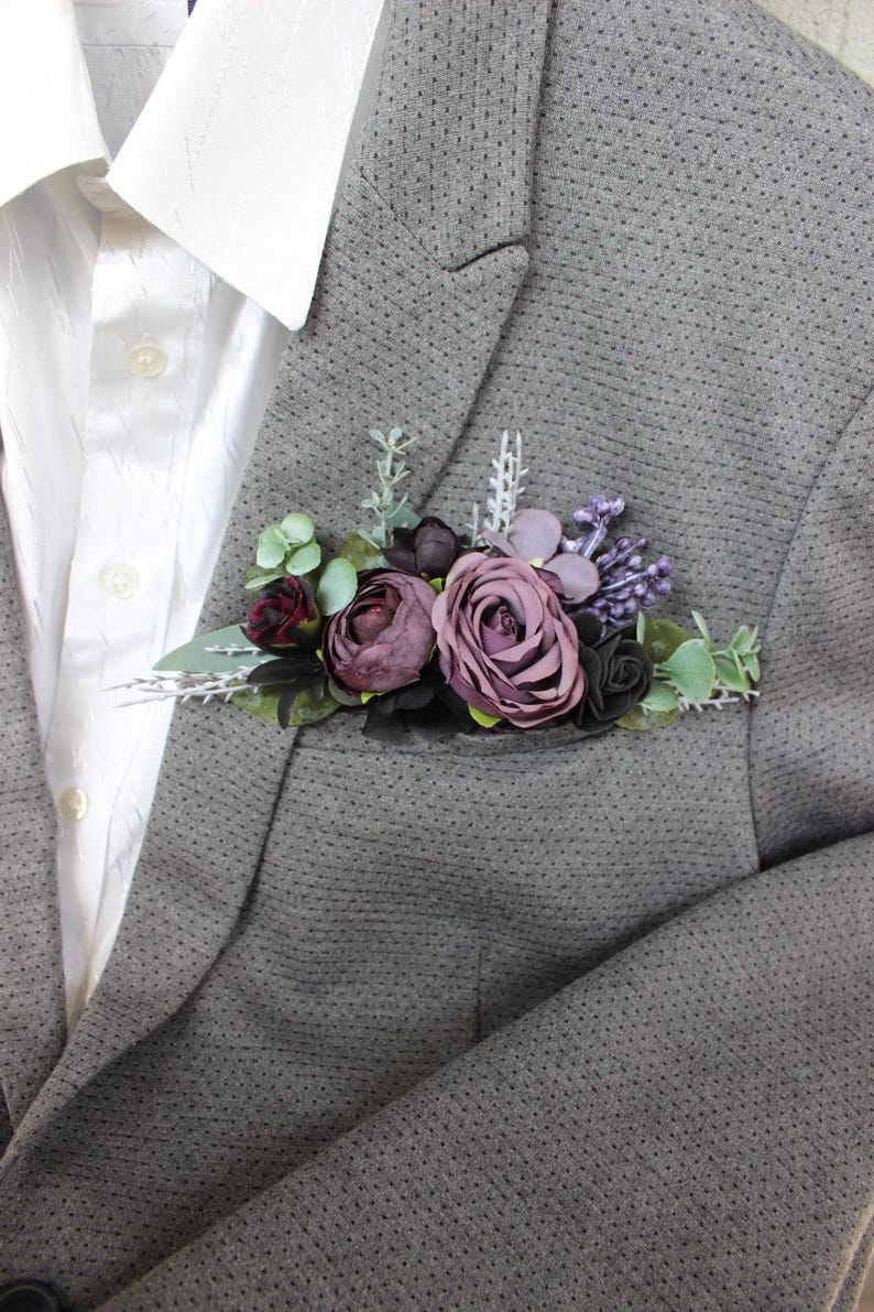 May include: A gray blazer with a floral pocket square featuring burgundy, purple, and green artificial flowers. The blazer is paired with a white dress shirt. The pocket square includes roses and other floral elements.