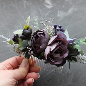 May include: A floral hair comb featuring dark purple and mauve roses, accented with green leaves and sprigs of white flowers. The comb is made of clear plastic and is designed for bridal or special occasion hairstyles.