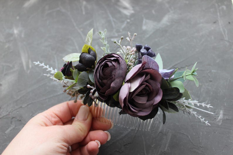 May include: A floral hair comb featuring dark purple and mauve roses, accented with green leaves and sprigs of white flowers. The comb is made of clear plastic and is designed for bridal or special occasion hairstyles.