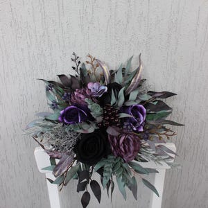 May include: A dark, moody floral arrangement featuring black and purple roses, accented with deep green and silver foliage. The bouquet is arranged on a white chair, creating a striking contrast. The overall theme is gothic and elegant.