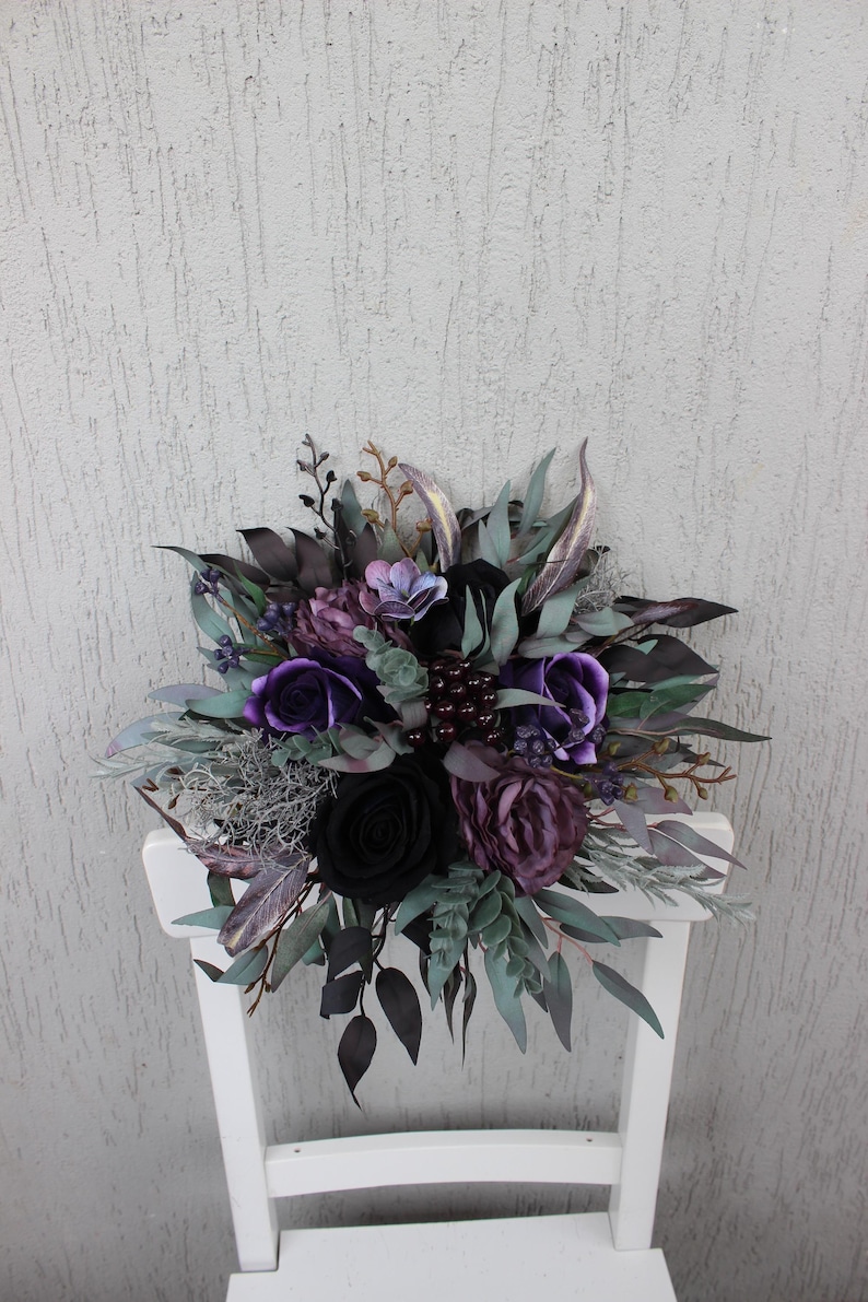 May include: A dark, moody floral arrangement featuring black and purple roses, accented with deep green and silver foliage. The bouquet is arranged on a white chair, creating a striking contrast. The overall theme is gothic and elegant.