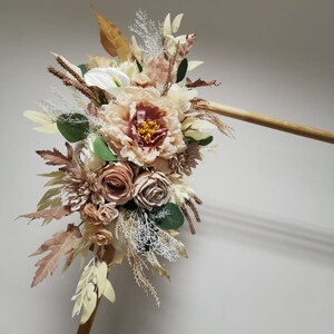 Puede incluir: Un ramo de flores artificiales en tonos marrones, beige y blancos. El ramo incluye rosas, peon&iacute;as y otros elementos florales. El ramo est&aacute; sujeto a una varilla de madera.