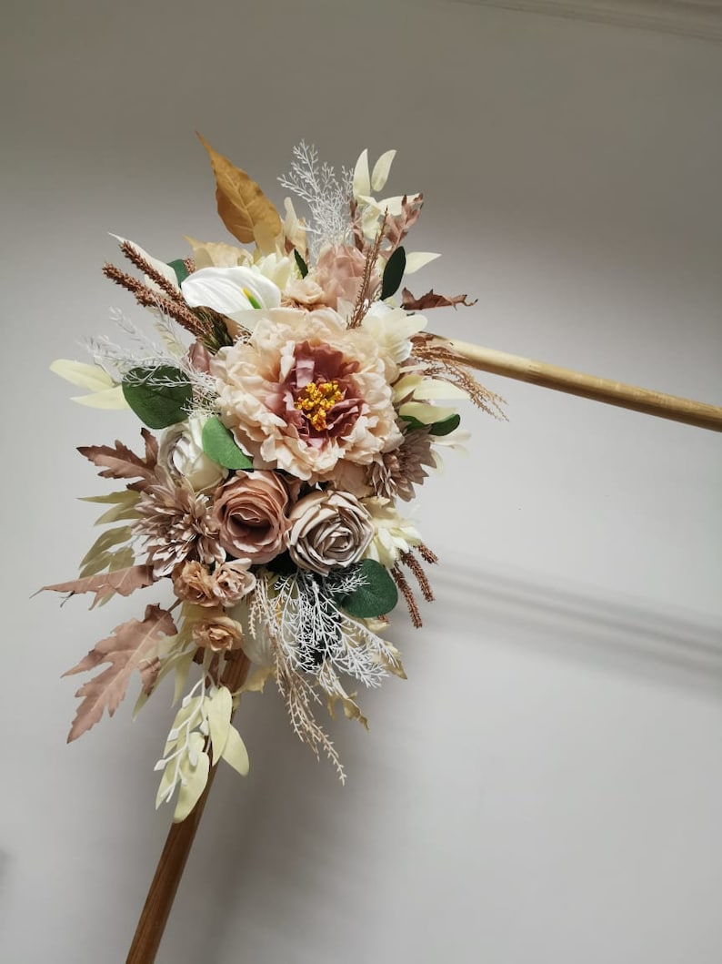 Puede incluir: Un ramo de flores artificiales en tonos marrones, beige y blancos. El ramo incluye rosas, peon&iacute;as y otros elementos florales. El ramo est&aacute; sujeto a una varilla de madera.