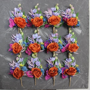 May include: Twelve orange flower boutonnieres with blue, purple, and green foliage. Each boutonniere has a single orange flower with a cluster of smaller flowers and leaves.