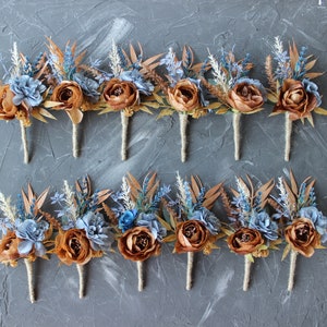May include: Ten boutonnieres featuring brown, blue, and tan artificial flowers and foliage. Each has a brown peony-like flower, blue and tan accents, and twine-wrapped stems. A rustic floral arrangement.