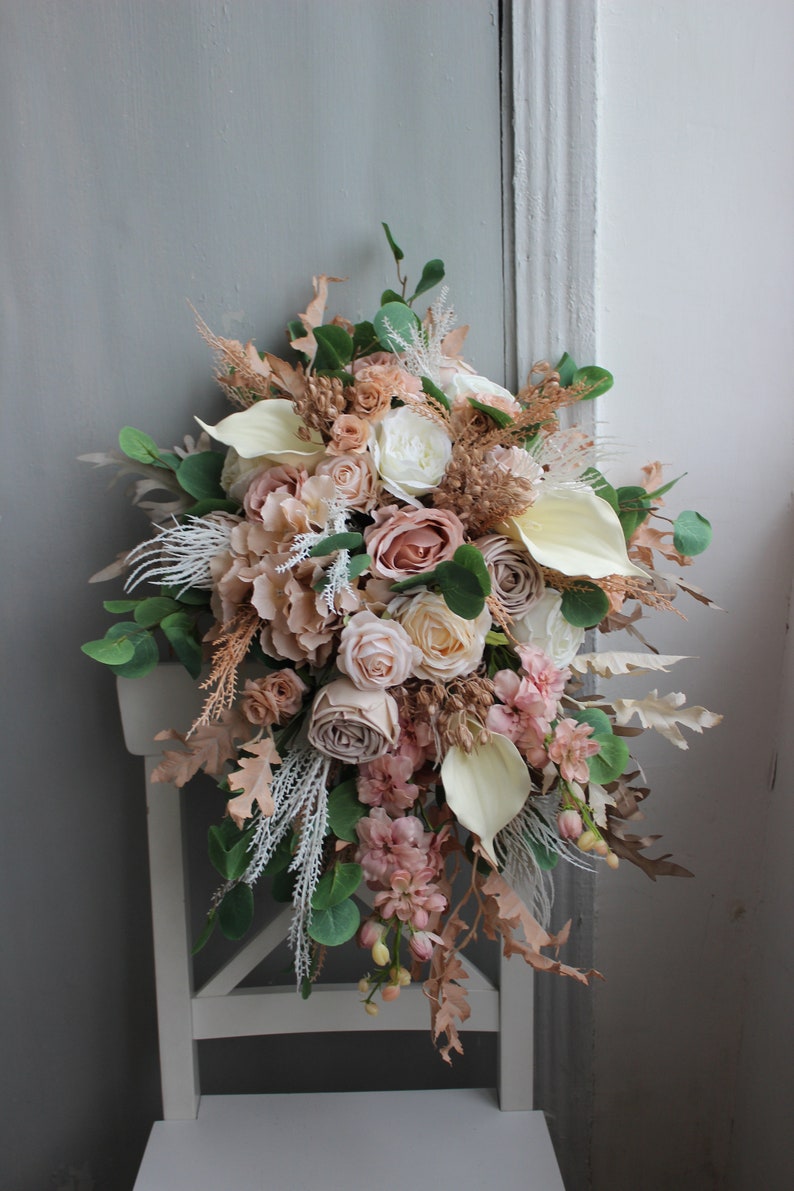 Puede incluir: Un ramo de novia en cascada con rosas de color rosa claro, blancas y crema, eucalipto y hierbas secas. El ramo est&aacute; dispuesto en forma de l&aacute;grima y es perfecto para una boda r&uacute;stica o bohemia.