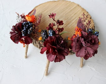 May include: Three burgundy and orange floral boutonnieres with blue berries and greenery.