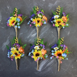 Op de afbeelding: Zes bloemenboutonnières met een zonnebloem als middelpunt. De boutonnières hebben gele, oranje, blauwe en paarse bloemen met groene bladeren. De stelen zijn omwikkeld met touw. De achtergrond is een donkergrijze ondergrond.