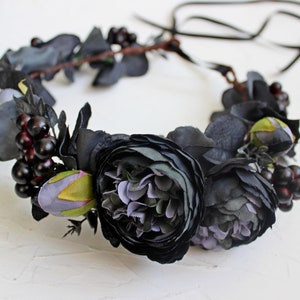 May include: A black floral crown with dark purple and green flowers and black berries. The crown is made of artificial flowers and is tied with a black ribbon.