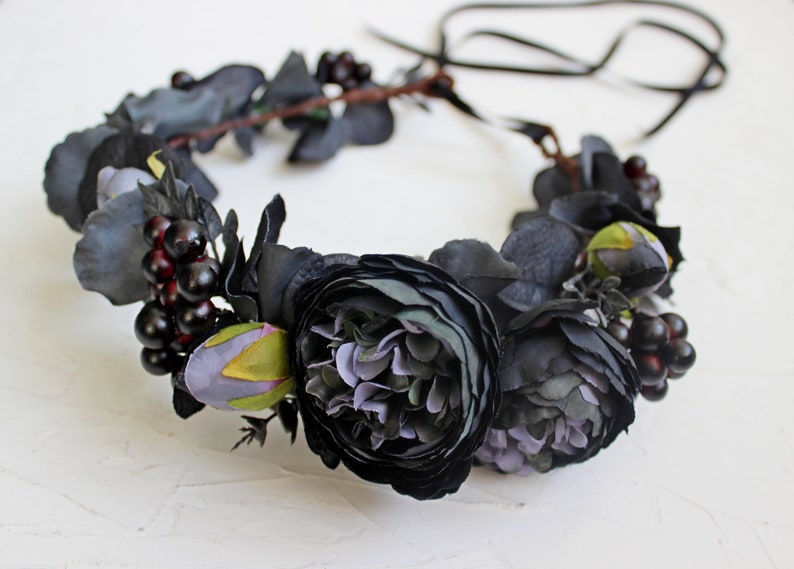 May include: A black floral crown with dark purple and green flowers and black berries. The crown is made of artificial flowers and is tied with a black ribbon.