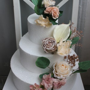 Puede incluir: Un maniqu&iacute; de pastel blanco de tres niveles decorado con flores artificiales de color rosa, crema y beige. El pastel est&aacute; adornado con una variedad de flores, incluyendo rosas, lirios calla y ramitas de vegetaci&oacute;n.