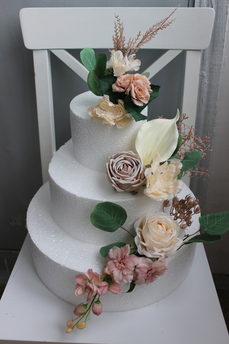 Puede incluir: Un maniqu&iacute; de pastel blanco de tres niveles decorado con flores artificiales de color rosa, crema y beige. El pastel est&aacute; adornado con una variedad de flores, incluyendo rosas, lirios calla y ramitas de vegetaci&oacute;n.