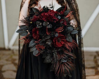 May include: A cascading floral bouquet featuring deep burgundy and black artificial flowers, including roses and anemones, accented with dark foliage, feathery plumes, and black leaves, held against a black dress with a black lace overlay.