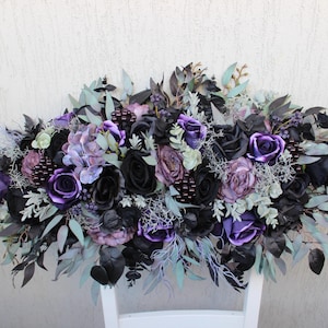 May include: A floral arrangement featuring a mix of black, purple, and lavender roses, hydrangeas, and other greenery. The arrangement is set against a white background and is suitable for weddings or home decor.
