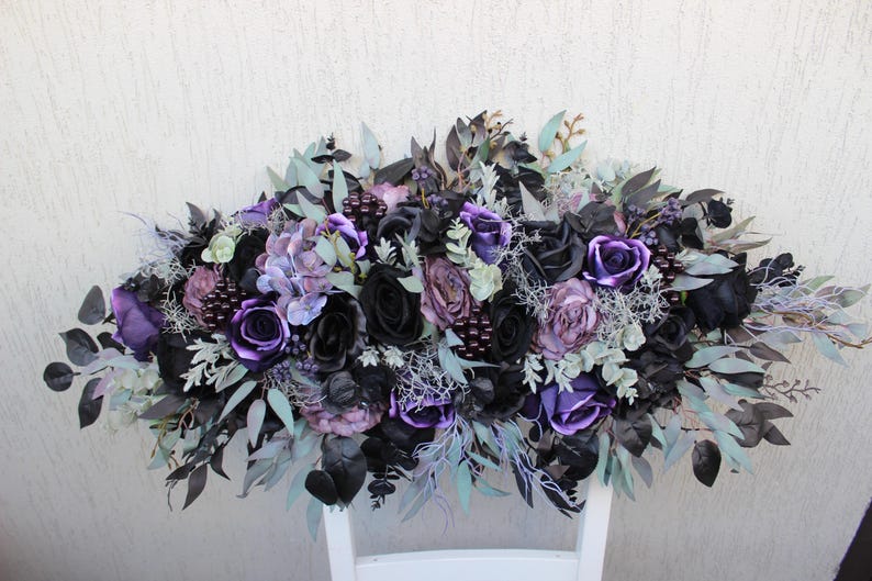 May include: A floral arrangement featuring a mix of black, purple, and lavender roses, hydrangeas, and other greenery. The arrangement is set against a white background and is suitable for weddings or home decor.