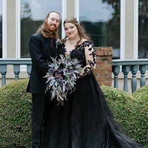 May include: A couple poses for a photo. The woman wears a black wedding gown with lace sleeves and a plunging neckline. She holds a bouquet of purple and green flowers. The man wears a black suit with a purple tie.