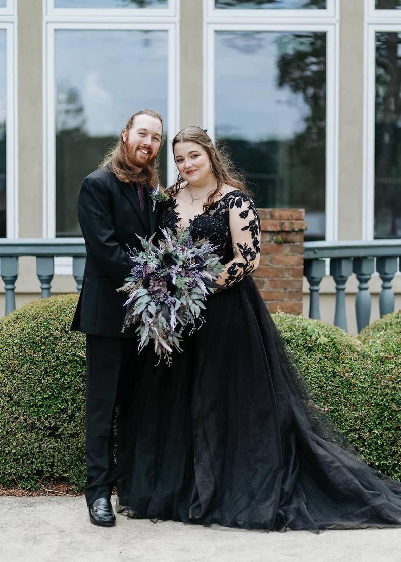 May include: A couple poses for a photo. The woman wears a black wedding gown with lace sleeves and a plunging neckline. She holds a bouquet of purple and green flowers. The man wears a black suit with a purple tie.