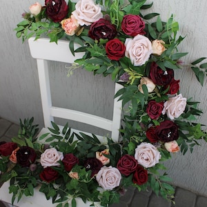 Puede incluir: Una silla de madera blanca adornada con un arreglo floral. El arreglo presenta rosas burdeos, rojas y rubor, con vegetación verde. Las flores caen en cascada sobre la silla, creando una exhibición decorativa.