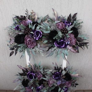 May include: Arrangement of artificial flowers in shades of purple and black, including roses and other blooms, with green and silver accents. The floral arrangement is displayed on a white surface.