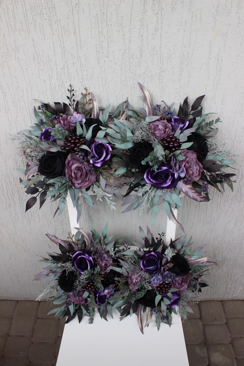 May include: Arrangement of artificial flowers in shades of purple and black, including roses and other blooms, with green and silver accents. The floral arrangement is displayed on a white surface.