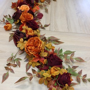 Op de afbeelding: Een bloemen tafelloper met kunstbloemen en bladeren in herfsttinten. De compositie bevat bordeauxrode rozen, oranje pioenrozen en bladeren in groen, geel en rood. De loper ligt op een licht houten oppervlak.