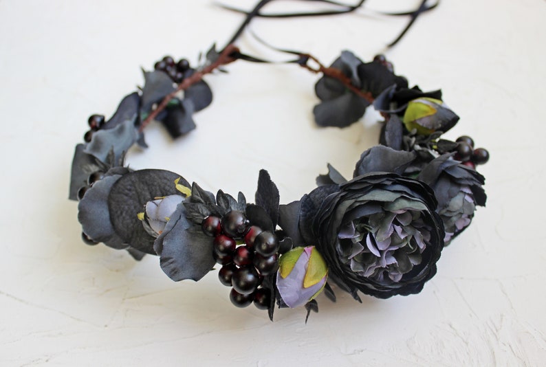 May include: A black floral crown with dark purple berries and black fabric flowers. The crown is made with a brown wire base and has black ribbon ties.