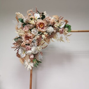 Puede incluir: Un arco de madera decorado con un arreglo floral de flores secas en tonos crema, beige y rosa. Las flores incluyen rosas, peon&iacute;as y otras flores delicadas.