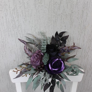 May include: A dark floral arrangement featuring deep purple and black roses, accented by green and gray foliage. The bouquet is arranged on a white chair, creating a striking contrast against the textured background.