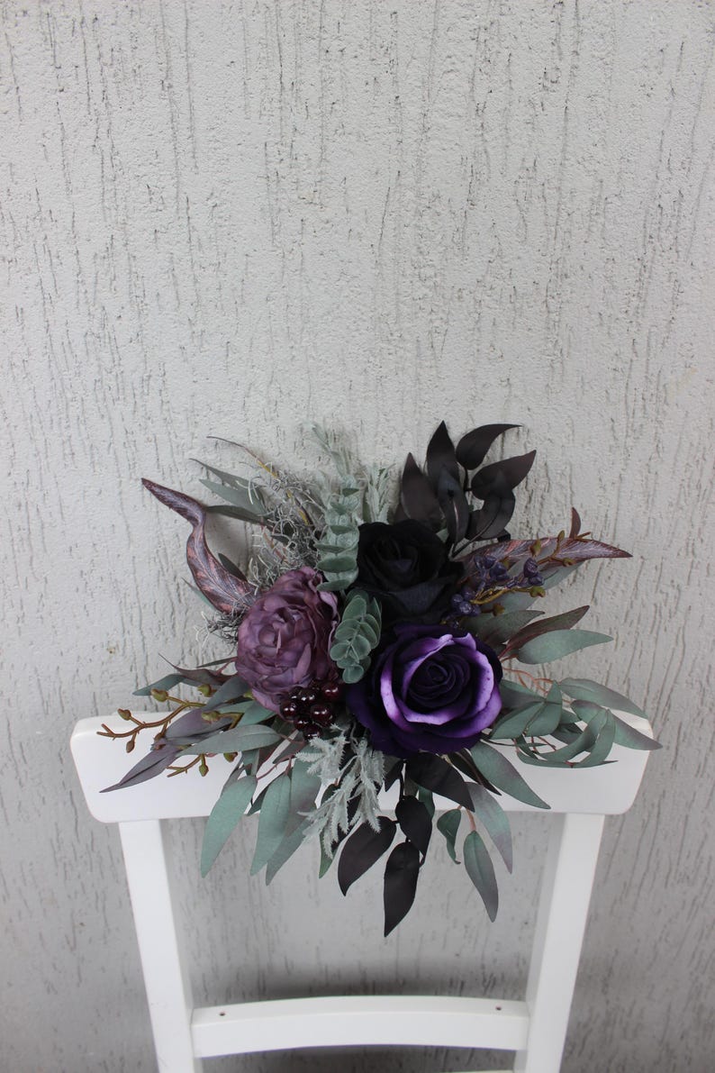 May include: A dark floral arrangement featuring deep purple and black roses, accented by green and gray foliage. The bouquet is arranged on a white chair, creating a striking contrast against the textured background.