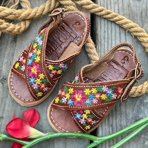 May include: A pair of brown leather sandals with colorful embroidered flowers. The sandals have a criss-cross strap design and a buckle closure.
