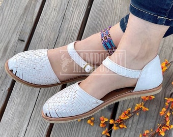 May include: White leather sandals with woven detailing and ankle straps. The sandals have a light brown sole and a small gold-colored buckle. The sandals are on a wooden surface. The brand name "RAMPOS MX" is visible in the top left corner.