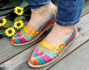 Puede incluir: Un par de sandalias de cuero tejidas de colores con un patrón amarillo, rosa, verde y azul. Las sandalias las lleva una persona que lleva pantalones vaqueros azules.