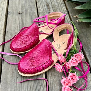 May include: A pair of pink leather sandals with intricate floral embroidery. The sandals have laces that tie around the ankles and are adorned with pink roses.