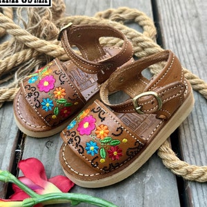 May include: A pair of brown leather sandals with colorful floral embroidery. The sandals have a buckle closure and a white stitched sole. The sandals are displayed on a wooden surface with a rope.