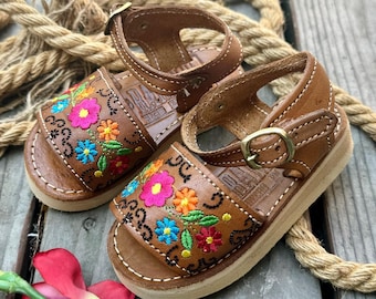 Può includere: Un paio di sandali in pelle marrone con ricami floreali colorati. I sandali hanno una chiusura con fibbia e una suola cucita bianca. I sandali sono esposti su una superficie di legno con una corda.