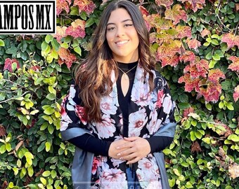 May include: A woman wearing a black top with a floral print shawl and blue jeans. She is standing in front of a wall of green and red leaves.