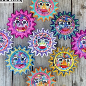 Puede incluir: Nueve decoraciones de pared con forma de sol pintadas a mano, coloridas y con diversos diseños y patrones. Cada sol tiene una cara diferente con características y colores únicos. Los soles están dispuestos en círculo sobre un fondo de madera.