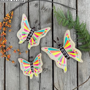 May include: Three colorful wooden butterflies with a rainbow pattern and brown accents. The butterflies are painted on a light brown wooden background.