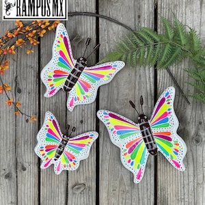 May include: Three colorful metal butterflies with white wings and rainbow accents. The butterflies are painted with black outlines and brown centers. The butterflies are arranged on a wooden surface.