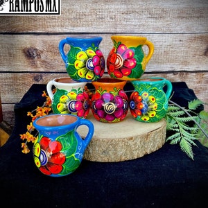 Op de afbeelding: Een verzameling van zes handbeschilderde keramische mokken. Elke mok heeft een uniek bloemenontwerp in verschillende kleuren, waaronder rood, geel, blauw en groen. De mokken worden gepresenteerd op een houten oppervlak.