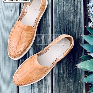 May include: A pair of tan leather sandals with a woven design. The sandals are on a wooden surface.