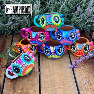 May include: A collection of colorful, hand-painted ceramic mugs with whimsical, owl-like faces. Each mug features a unique combination of bright colors, including blue, pink, orange, and yellow, with intricate floral and geometric patterns. The mugs are displayed on a wooden surface.