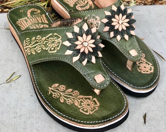 Pode incluir: Um par de sandálias de couro verde com bordado floral e um enfeite de flor preta e rosa. As sandálias têm um detalhe de costura branca ao redor da sola.