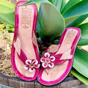 May include: A pair of pink leather sandals with floral embellishments. The sandals have a brown leather strap with the text "RANDPOS" and "MADE IN MEXICO" on them.