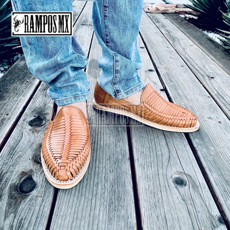 May include: A pair of tan leather sandals with a woven design. The sandals are being worn by someone wearing blue jeans. The sandals are on a wooden surface.