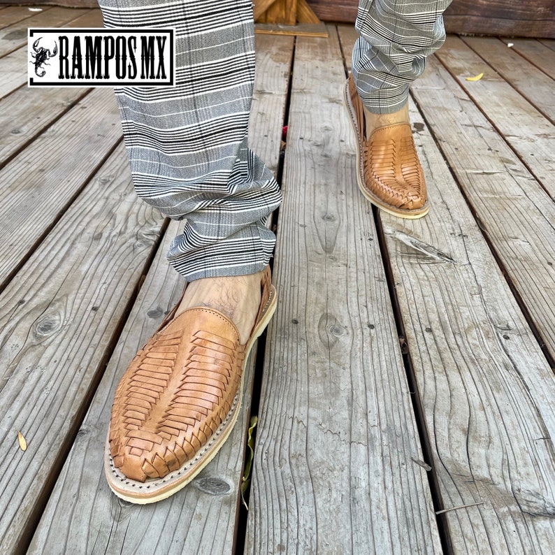 May include: A pair of brown leather sandals with woven straps. The sandals are being worn by a person wearing grey plaid trousers. The sandals are on a wooden deck.