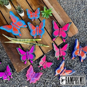 May include: A collection of colorful butterfly-shaped ceramic wall decor. The butterflies are painted in various shades of pink, blue, and purple. Some butterflies have black outlines, while others have white outlines. The butterflies are arranged on a wooden surface.