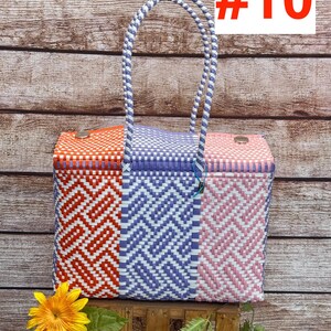Op de afbeelding: Een rechthoekige gevlochten plastic tas met een driekleurig geometrisch patroon in oranje, paars en roze met witte accenten. De tas heeft twee gevlochten handvatten en twee metalen sluitingen aan de bovenrand. Tekst RAMPOS MX en #10 zichtbaar.