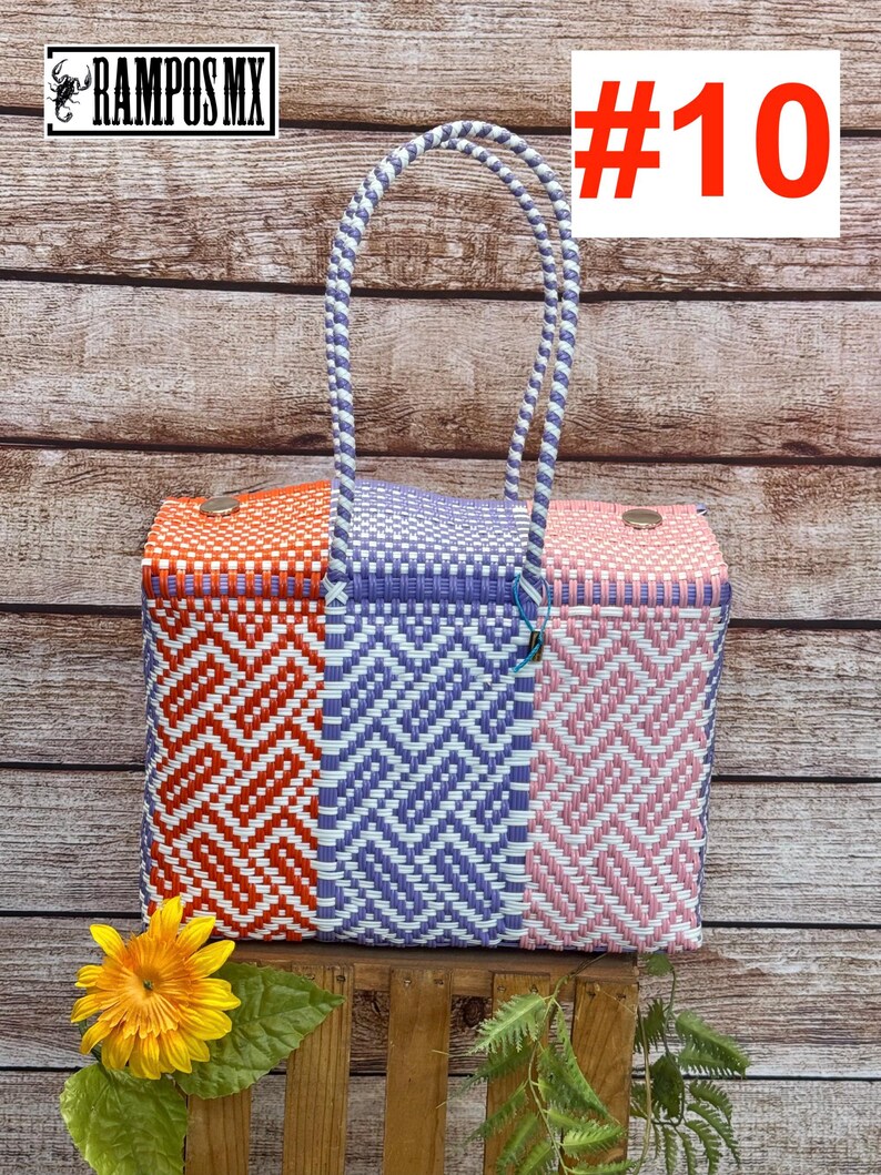 Op de afbeelding: Een rechthoekige gevlochten plastic tas met een driekleurig geometrisch patroon in oranje, paars en roze met witte accenten. De tas heeft twee gevlochten handvatten en twee metalen sluitingen aan de bovenrand. Tekst RAMPOS MX en #10 zichtbaar.